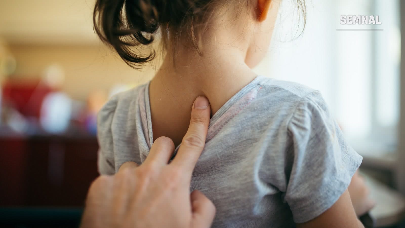 Fetiță văzută din spate, în tricou gri, în timp ce mâna unui adult îi atinge coloana vertebrală la baza gâtului, în timpul unui consult