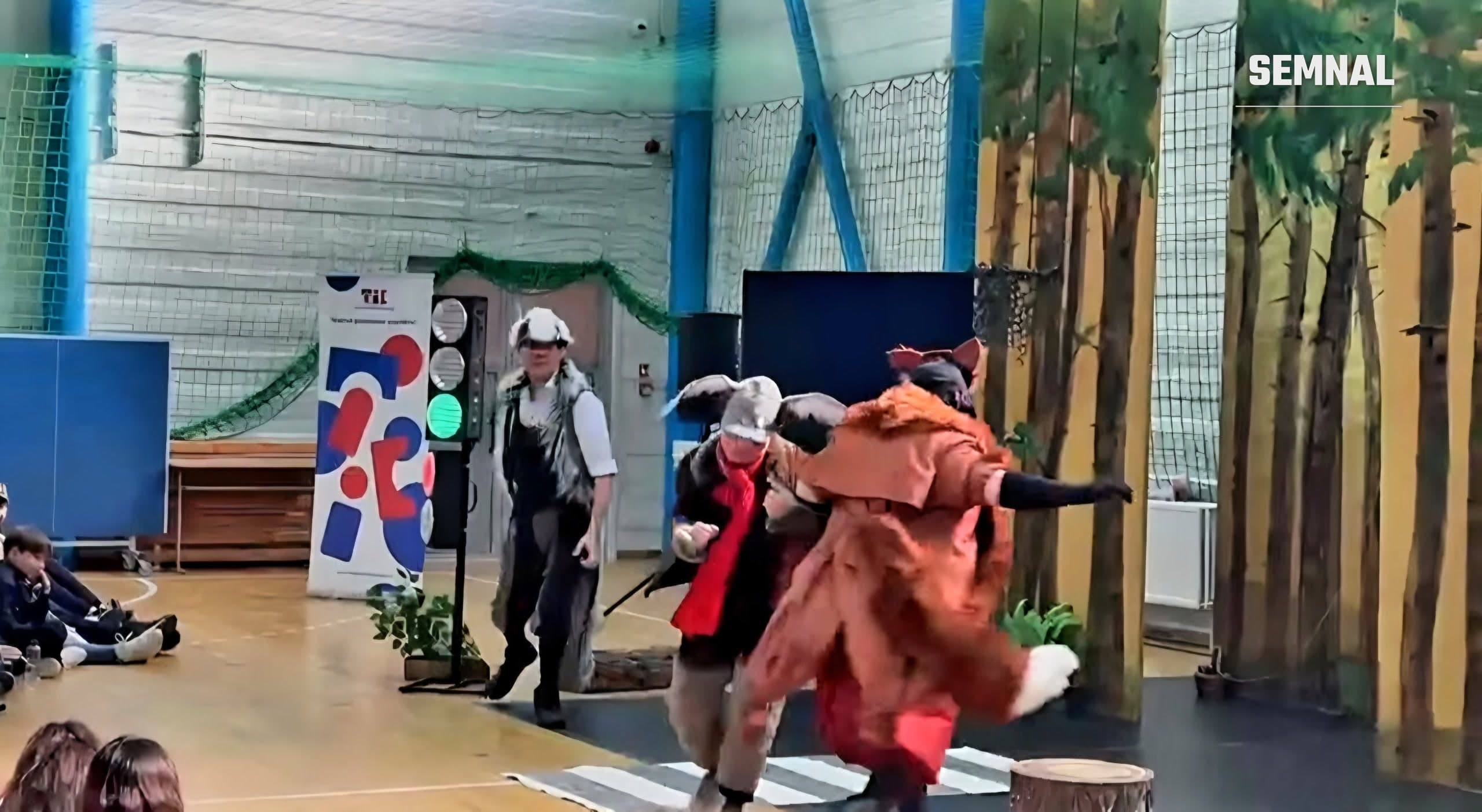 Actori costumați, printre care unul în costum de vulpe, pe o scenă amenajată într-o sală de sport, cu decor de pădure, semafor și trecere de pietoni; în stânga stau copii pe jos