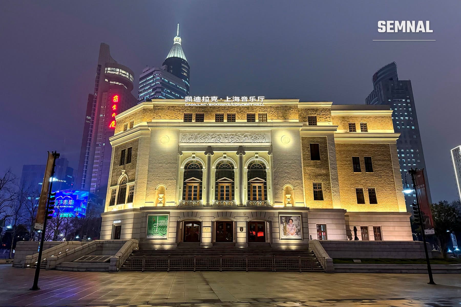 Fațada iluminată noaptea a Sălii de Concerte din Shanghai, clădire din 1930, cu blocuri turn în fundal