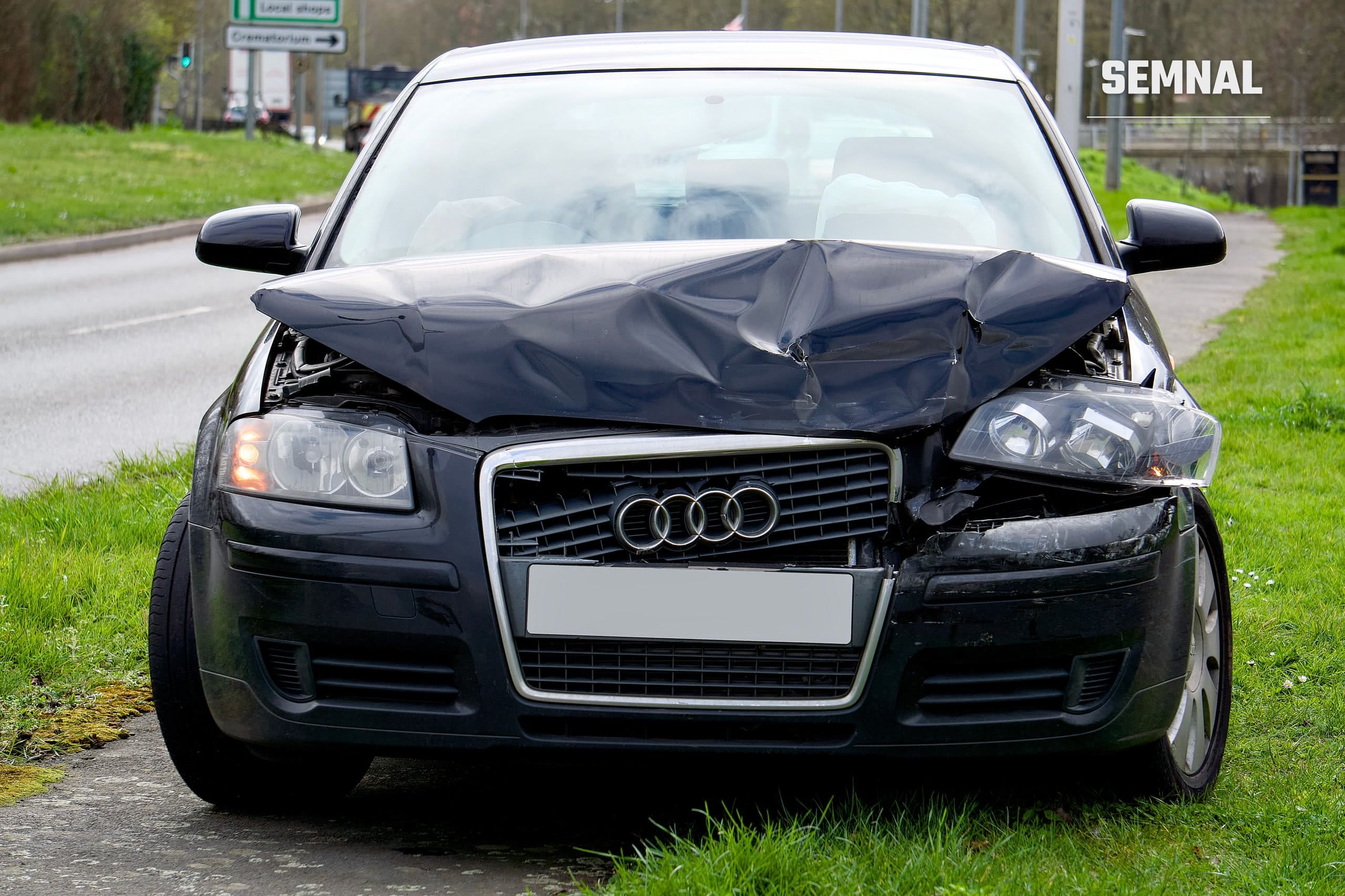 Mașină neagră cu avarii semnificative la partea frontală, pe marginea drumului, după un accident.