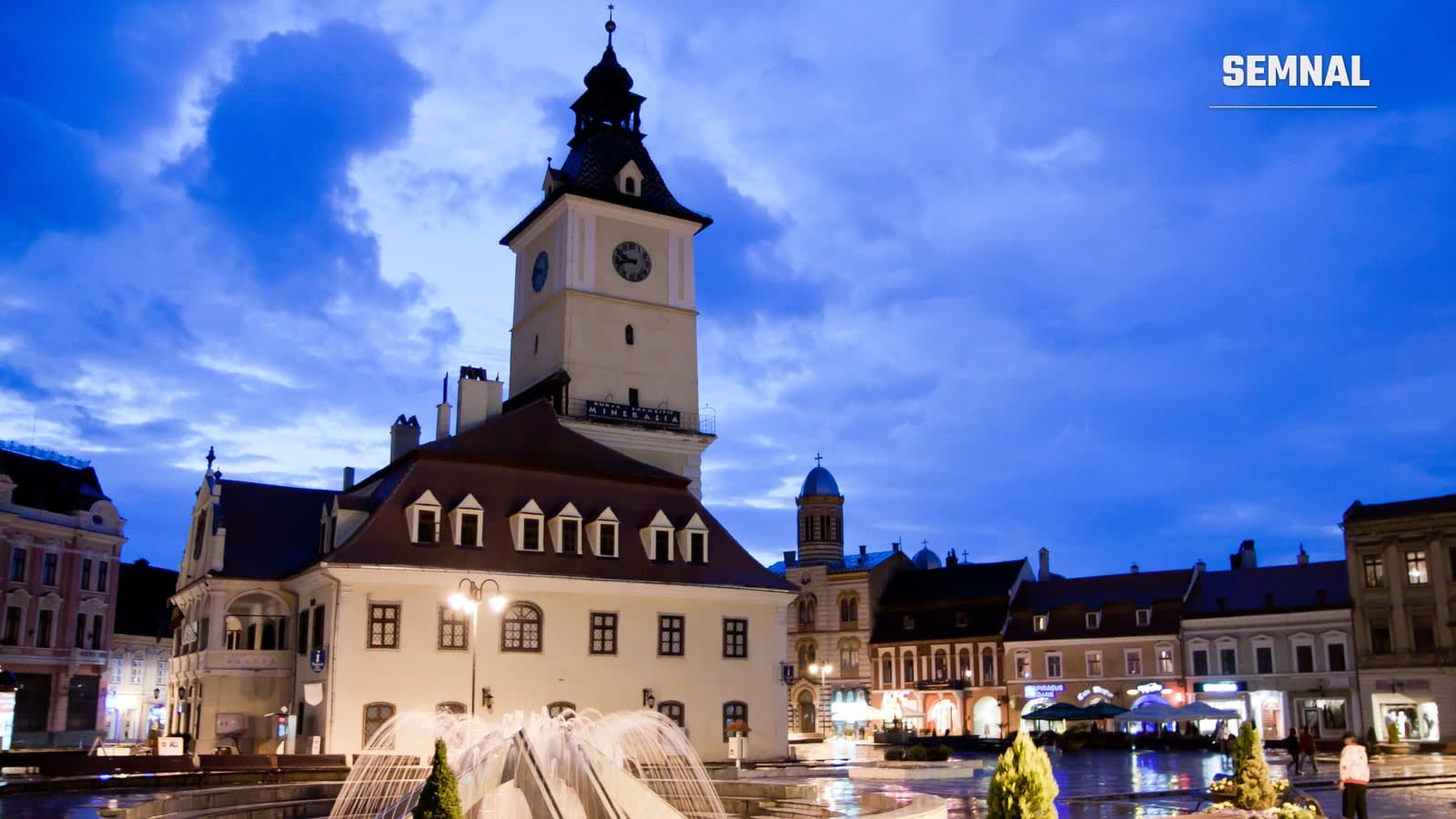 Piața Sfatului din Brașov seara, cu Casa Sfatului și turnul cu ceas, o fântână arteziană și bănci în prim-plan