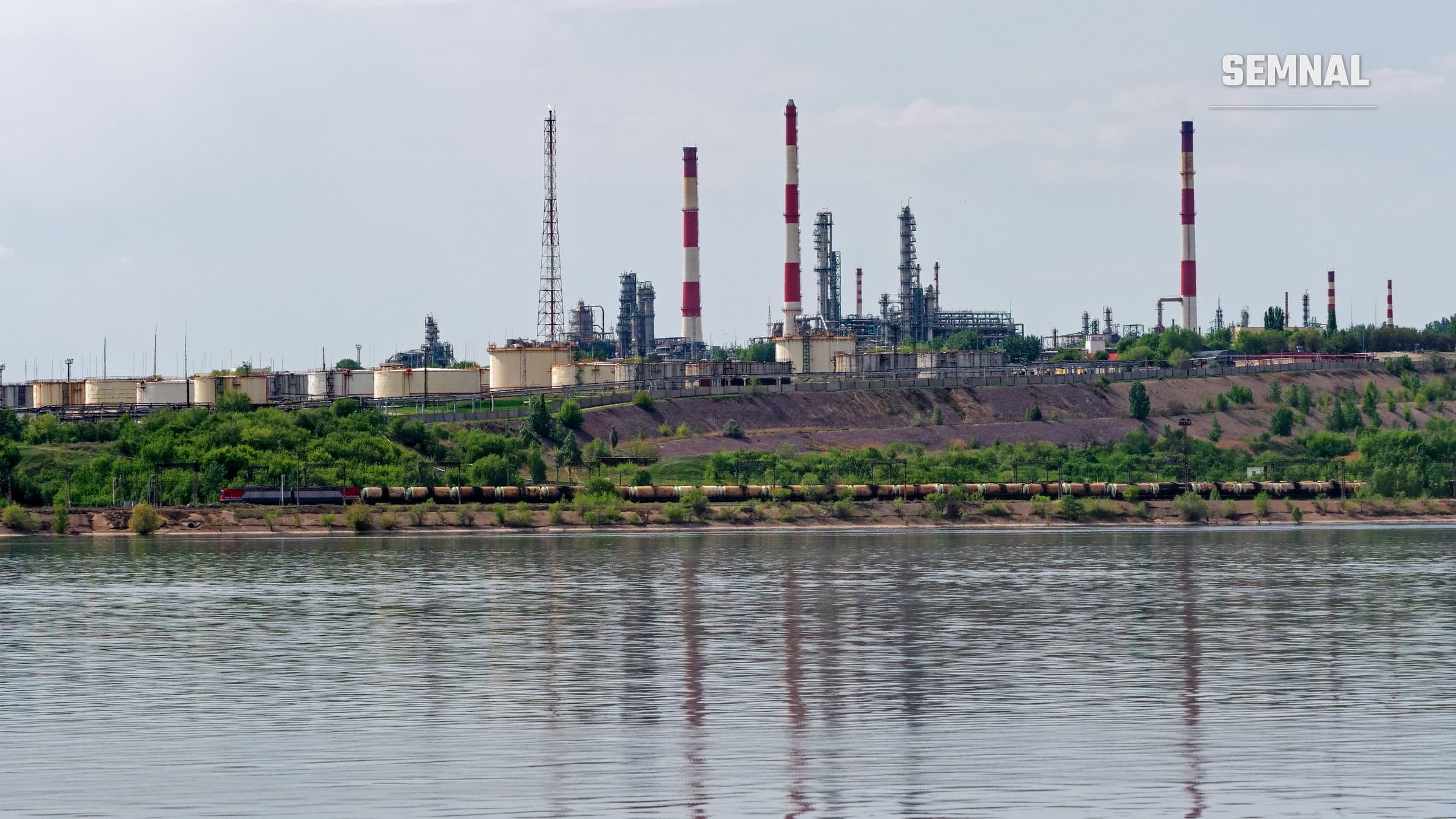 Rafinăria de petrol din Saratov, văzută de pe Volga: coșuri de fum alb-roșii, turnuri de distilare și rezervoare pe malul apei