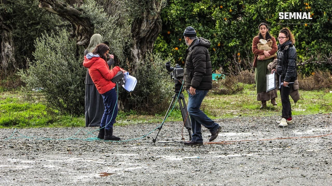 Echipă de filmare pe un platou în aer liber, cu o cameră pe tripod și actrițe în costume de epocă