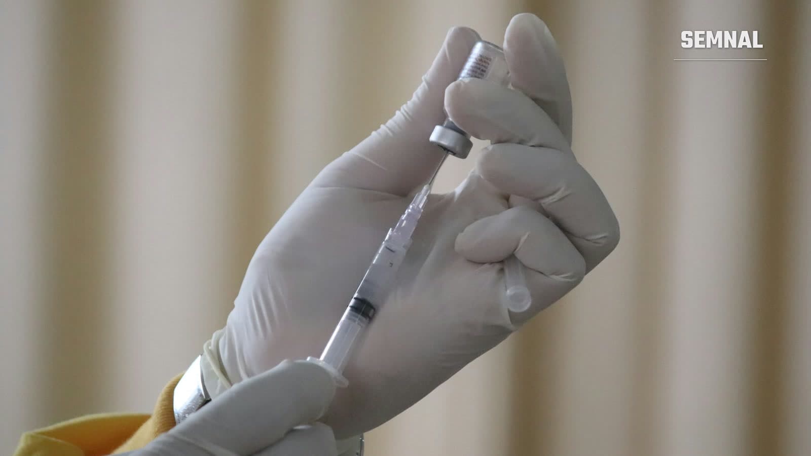 Mâini în mănuși albe de latex umplu o seringă dintr-o fiolă de vaccin, într-o încăpere cu draperii bej