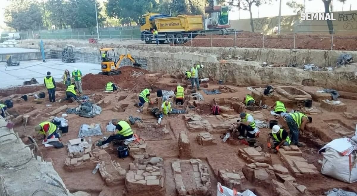 Arheologi în veste reflectorizante lucrează într-o excavație cu morminte din țigle și cărămizi, pe fundal un excavator și un camion