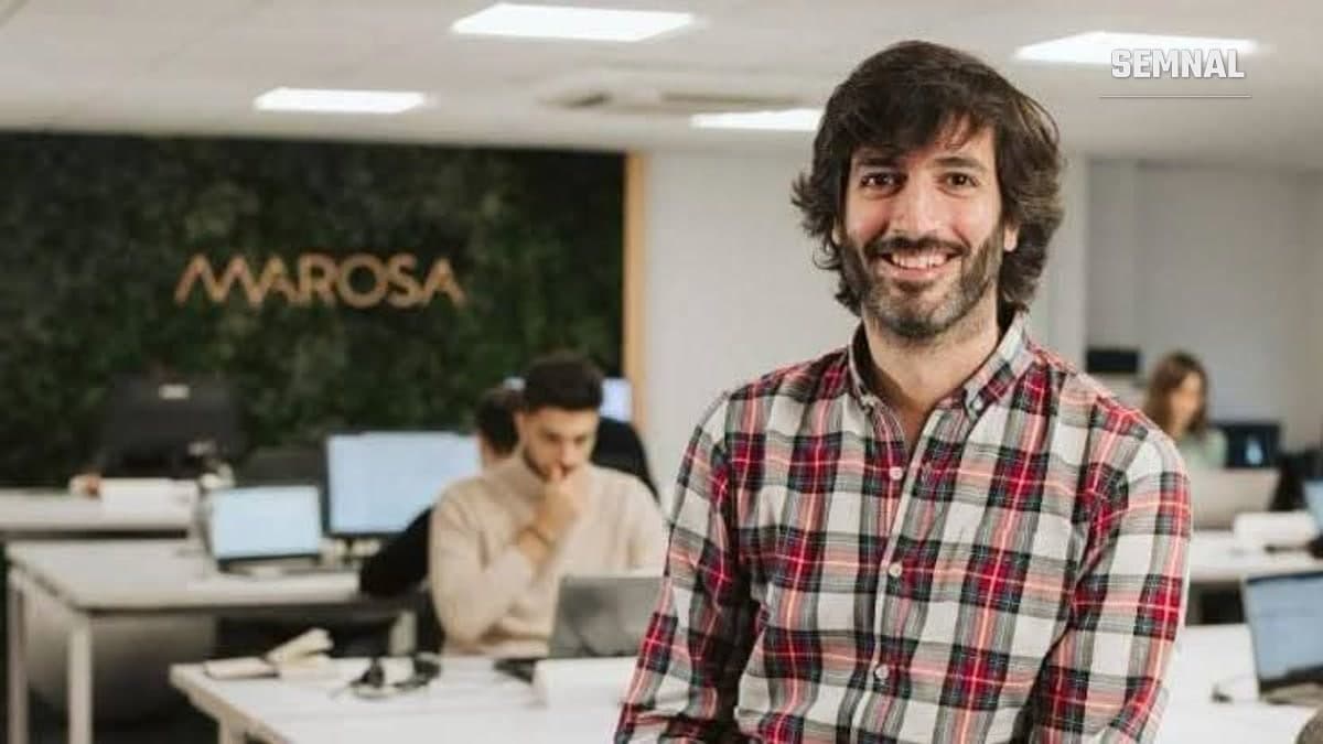 Bărbat zâmbitor, cu barbă și cămașă în carouri, într-un birou open space; în fundal, logoul Marosa pe un perete verde și colegi la calculatoare