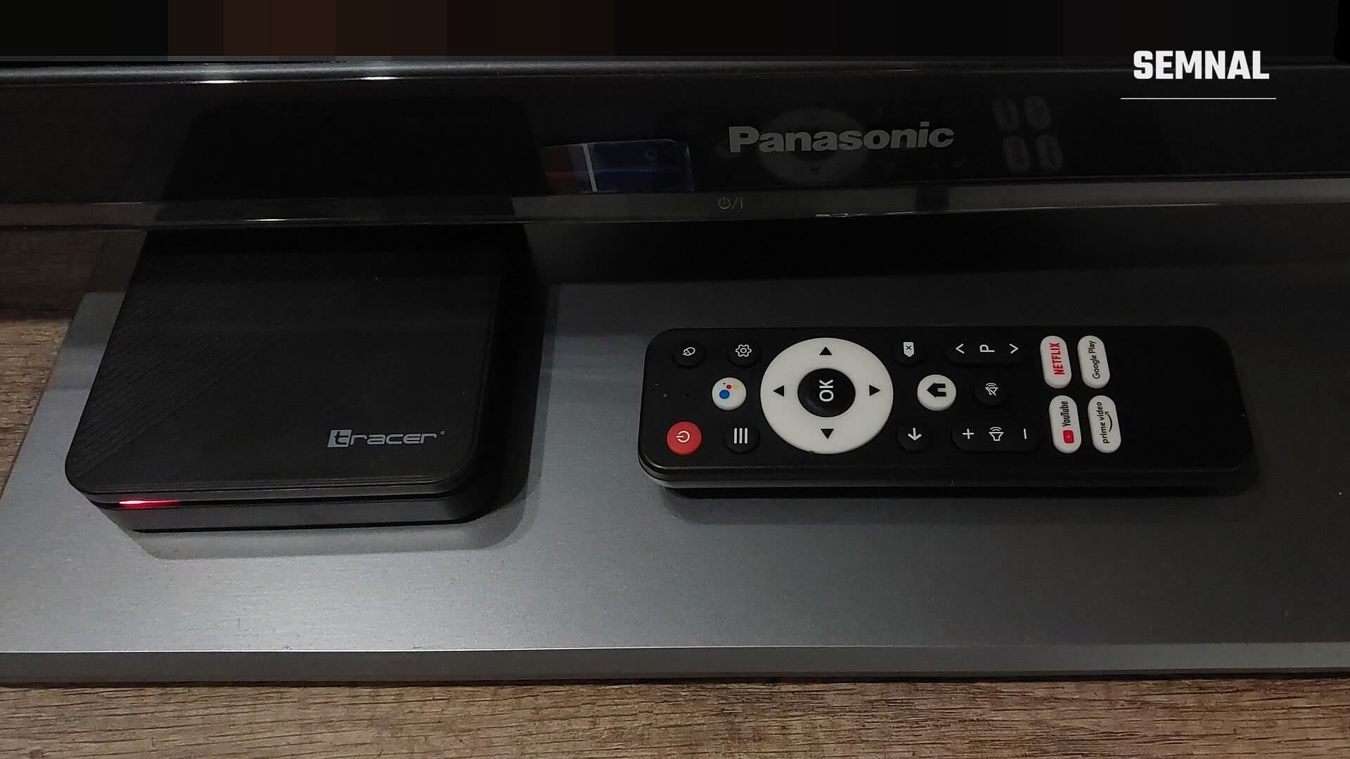 Cutie Android TV Tracer, neagră, cu telecomanda alăturată, așezată sub un televizor Panasonic
