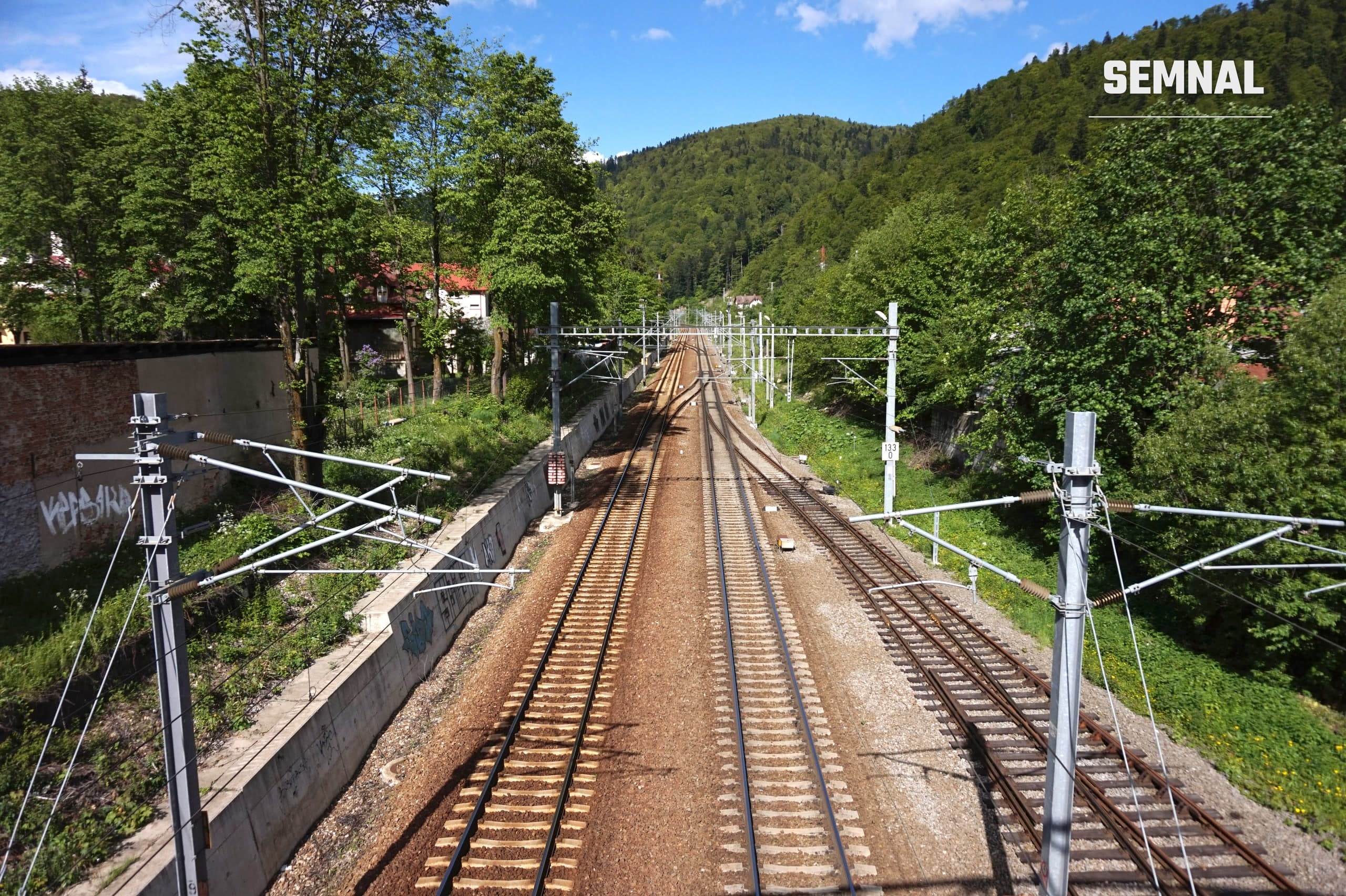 Linii de cale ferată electrificată la Bușteni, cu șine, traverse și piatră spartă, între dealuri împădurite. Foto: Tiia Monto / Wikimedia Commons, CC BY-SA 4.0