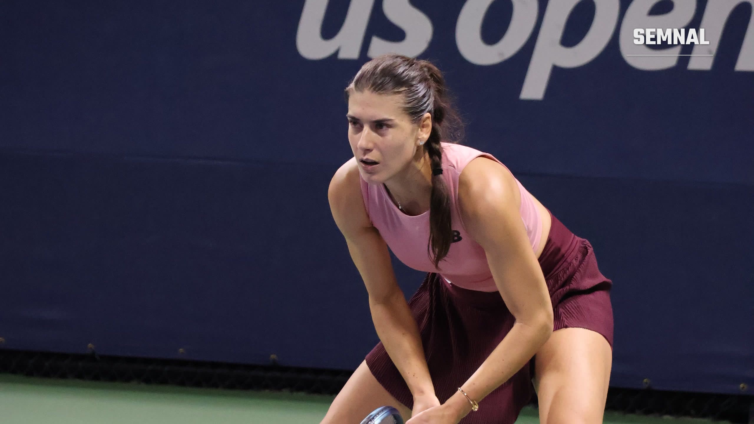 Sorana Cîrstea, în top roz și fustă vișinie, stă aplecată în poziție de așteptare, cu racheta în mână, în fața unui panou US Open