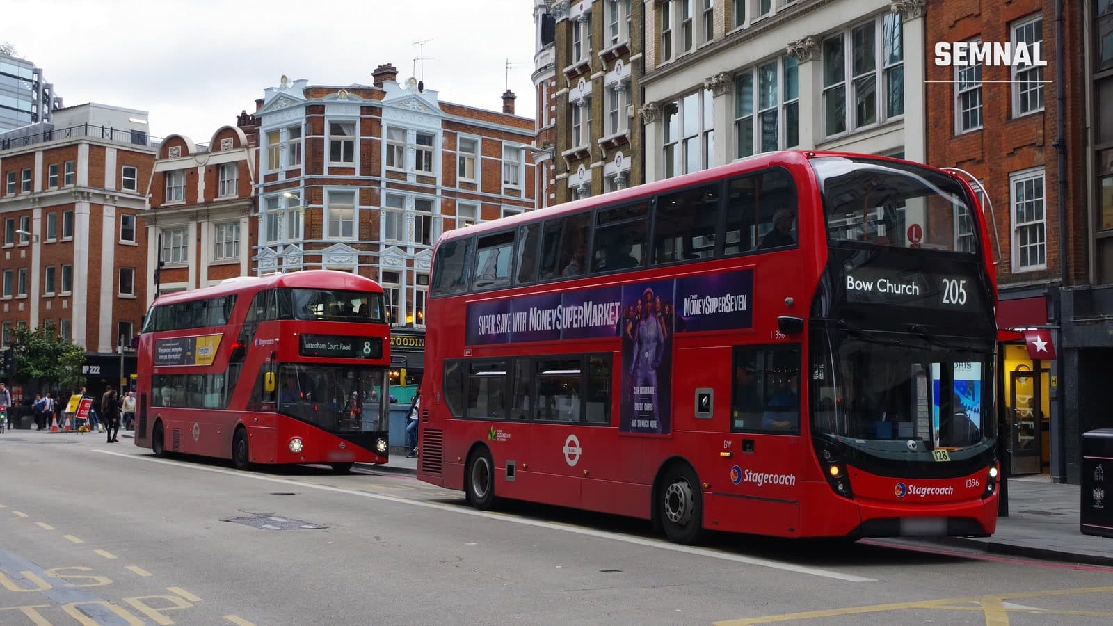 Două autobuze etajate roșii, pe rutele 205 și 8, circulând pe o stradă din Londra, între clădiri din cărămidă