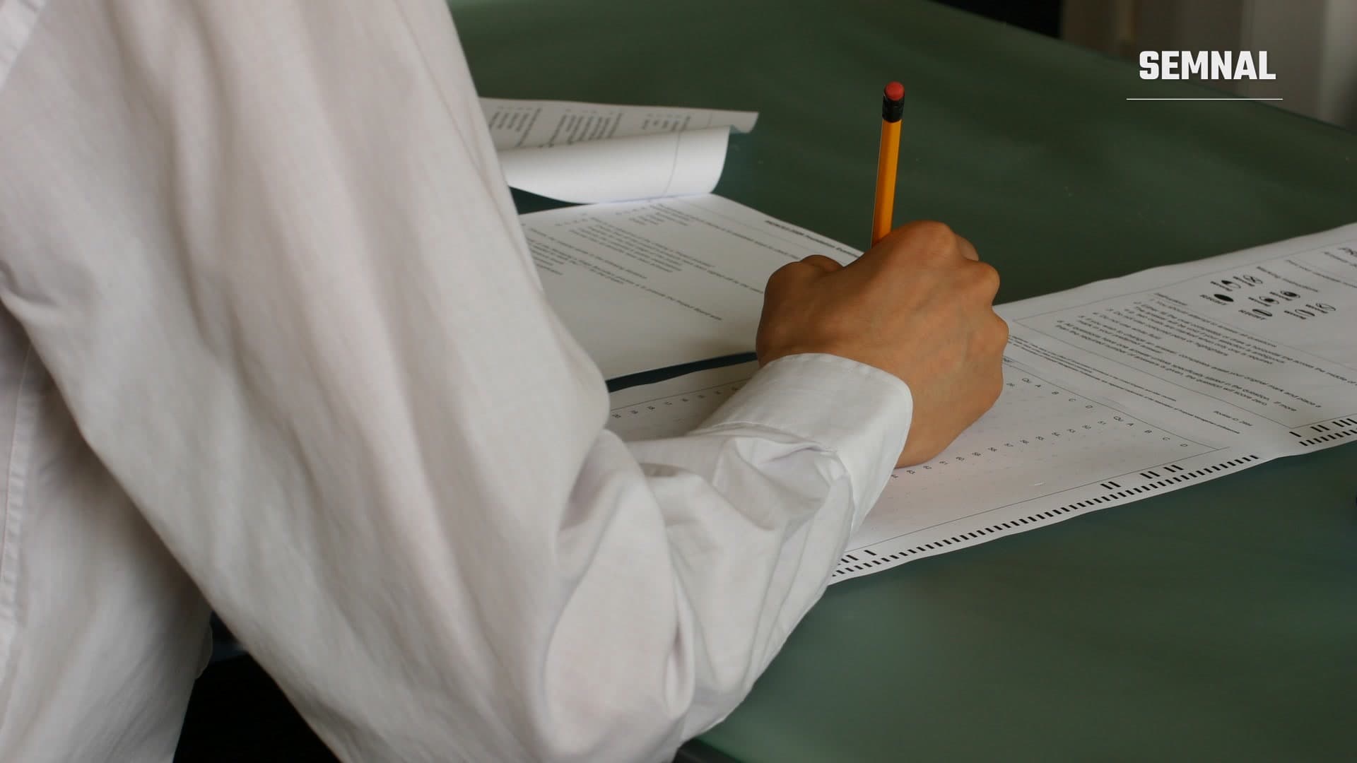 O mână care scrie cu un creion galben pe o foaie de examen cu răspunsuri de bifat, așezată pe un blat verde, într-o sală de clasă