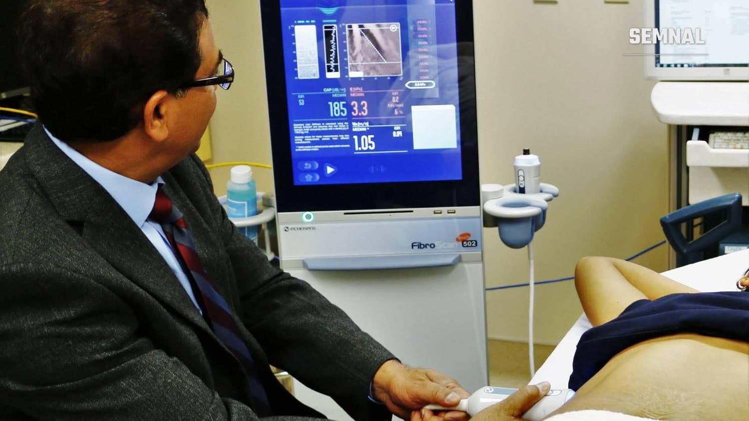 Medic în costum privește ecranul unui aparat FibroScan în timp ce ține sonda pe abdomenul unui pacient, în timpul unei elastografii hepatice