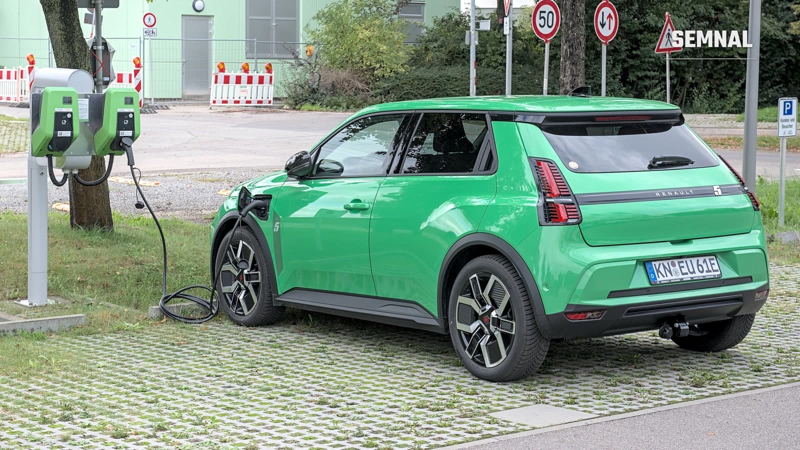 Renault 5 E-Tech Electric verde, văzut din lateral-spate, parcat pe o parcare cu dale înierbate și conectat prin cablu la o stație de încărcare cu două prize