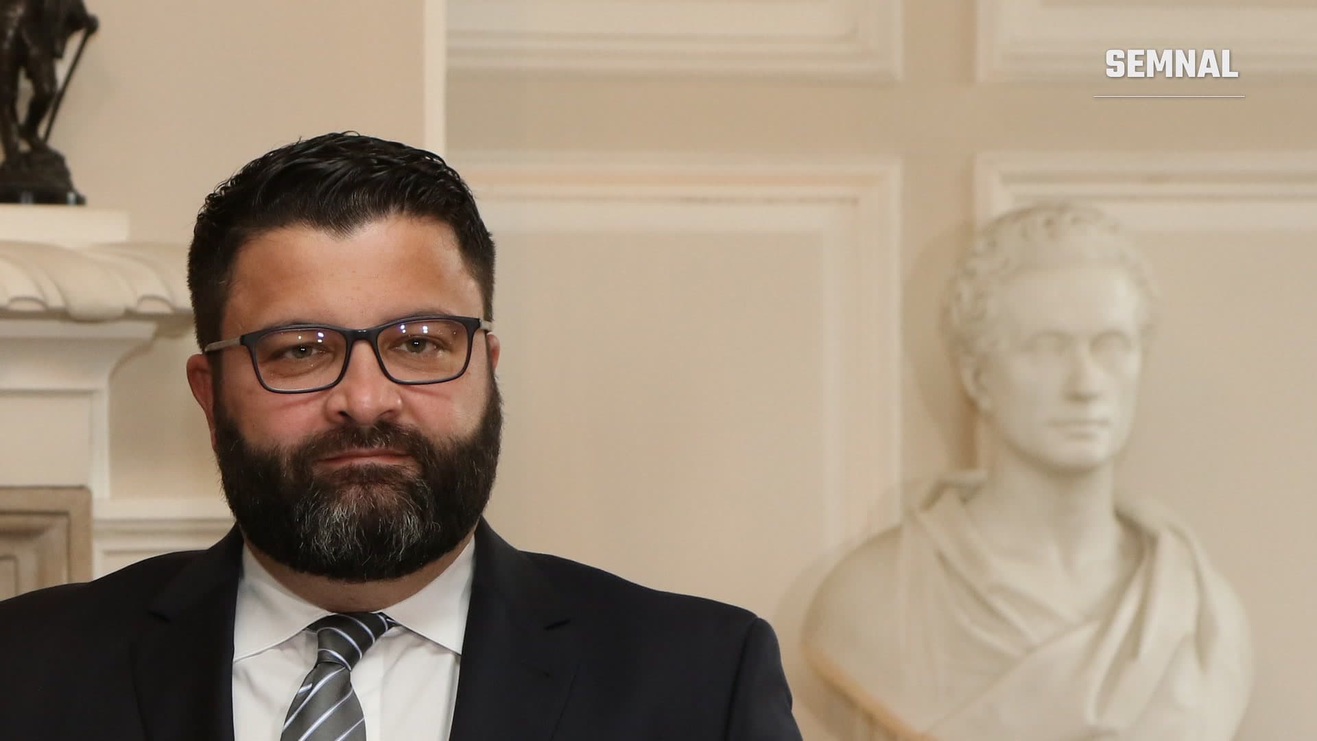Portret de arhivă (2018) al lui Dan Neculăescu, ministrul român de Externe: bărbat cu barbă și ochelari, în costum închis, lângă un bust de marmură.