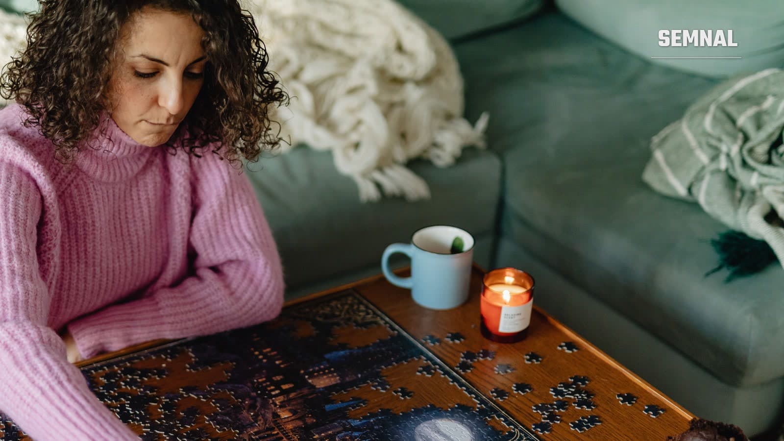 Femeie în pulover roz care asamblează un puzzle într-un living, lângă o ceașcă și o lumânare aprinsă (Foto: Kaboompics / Pexels)