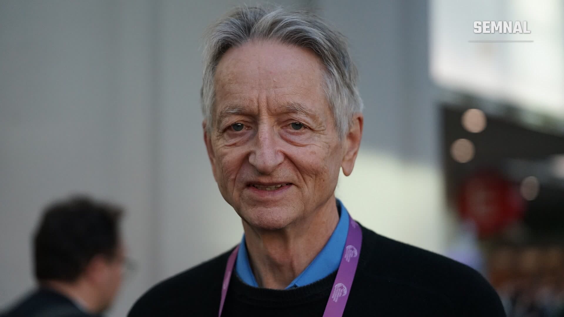 Geoffrey Hinton, în prim-plan, cu un șnur mov la gât, într-un spațiu de conferință la San Diego, în decembrie 2025 (Foto: Xuthoria / Wikimedia Commons, CC BY-SA 4.0)
