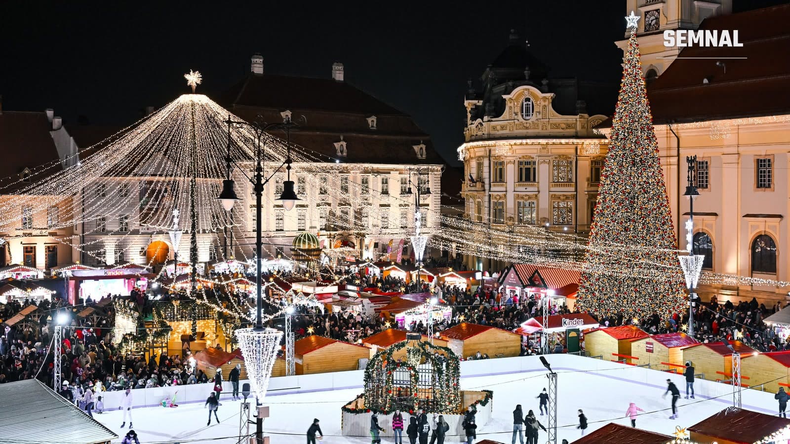 Piața Mare din Sibiu iluminată noaptea în timpul Târgului de Crăciun, cu brad uriaș, cupolă de luminițe, căsuțe din lemn și patinoar cu oameni care patinează