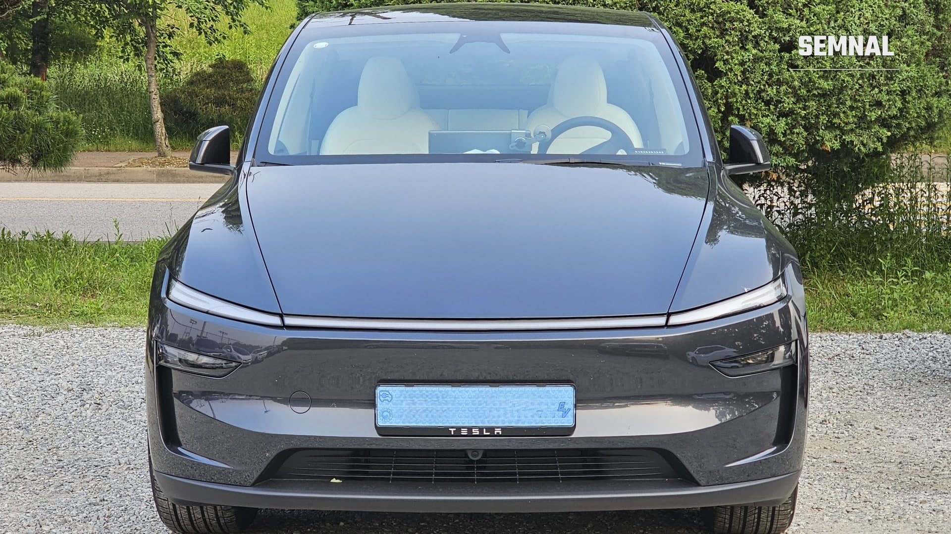 Tesla Model Y gri închis, văzut din față, parcat pe pietriș, cu bara luminoasă dintre faruri și sigla Tesla pe bara de protecție