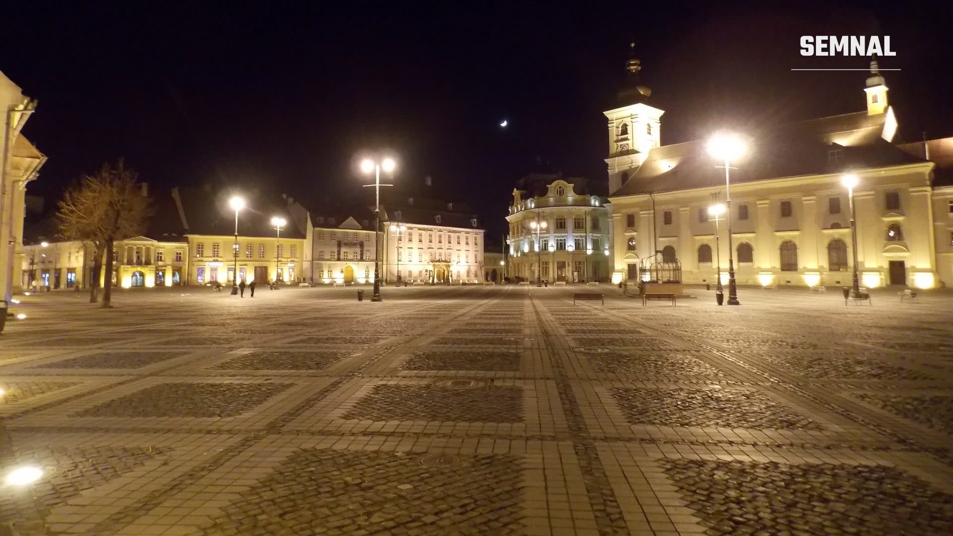 Piața Mare din Sibiu noaptea, goală, cu clădiri istorice luminate, felinare aprinse și o semilună pe cerul întunecat