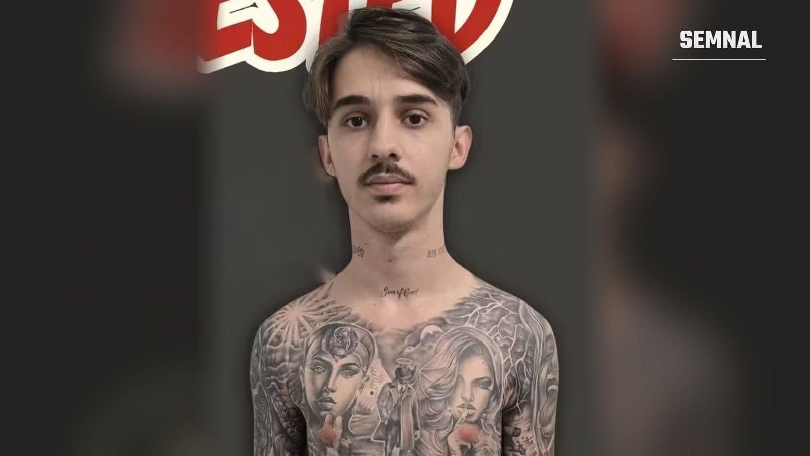 Tânăr român arestat de Border Patrol, cu mustață și tatuaje pe piept, fotografiat pe un fundal gri