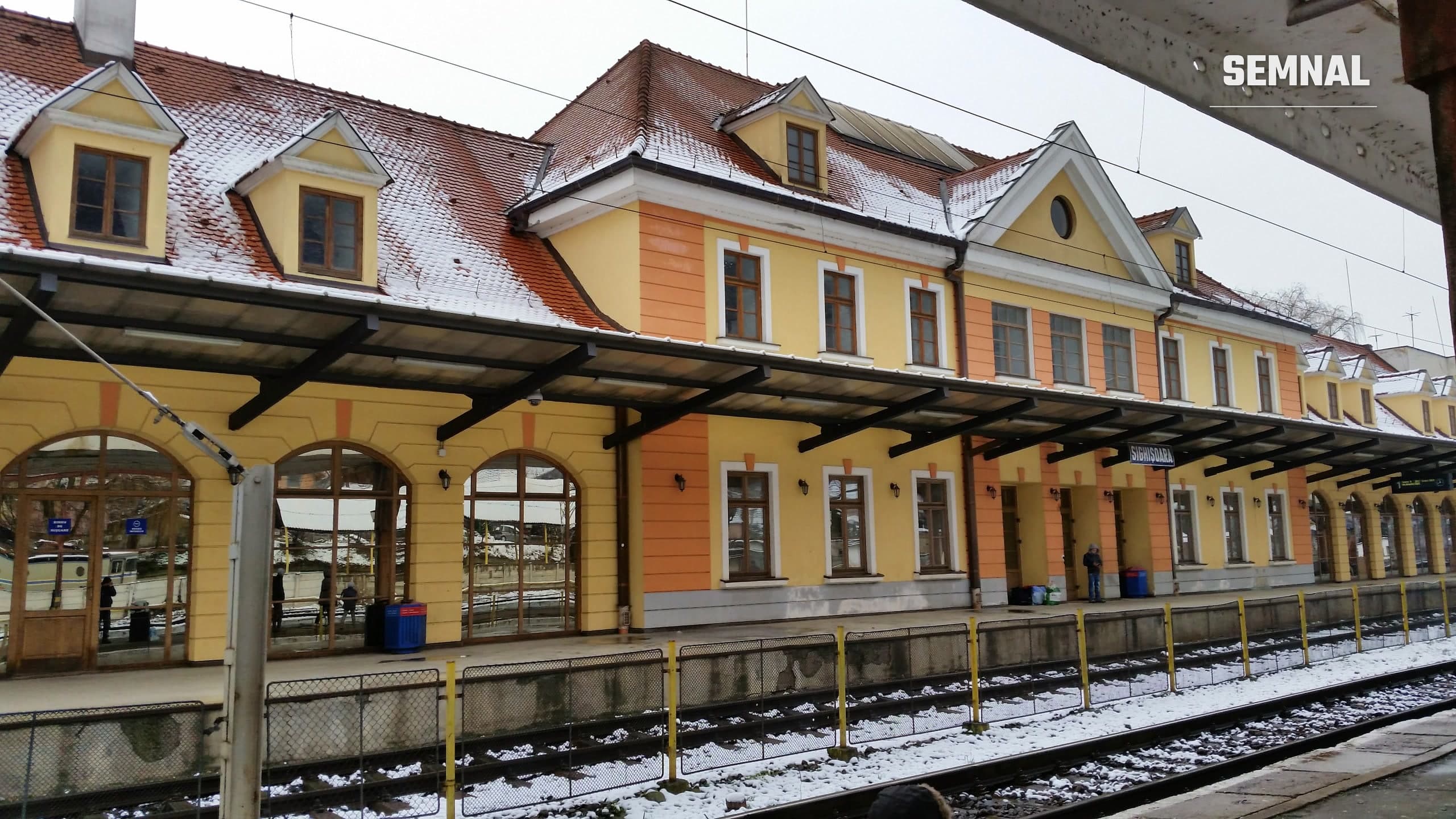 Clădirea gării din Sighișoara, cu fațadă galbenă și portocalie, acoperiș cu țiglă roșie acoperit de zăpadă și linii de cale ferată în prim-plan. Imagine ilustrativă din 2014. (Foto: PosifCool / Wikimedia Commons, CC BY-SA 4.0)