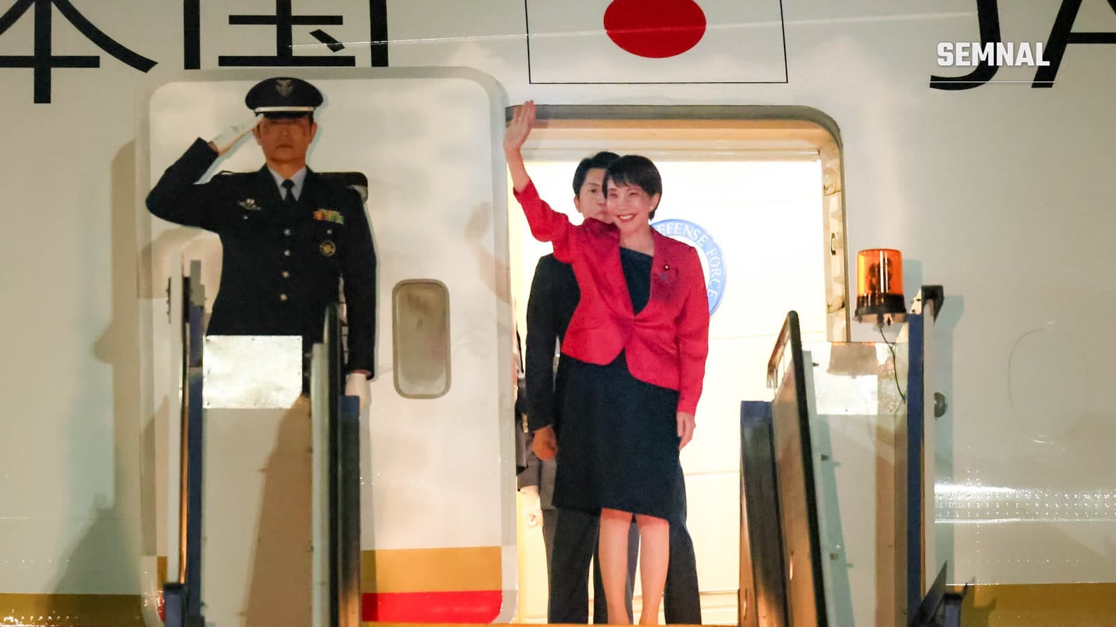 Premierul japonez Sanae Takaichi salută din ușa avionului guvernamental la sosirea în Australia, în mai 2026, lângă un militar care face onorul