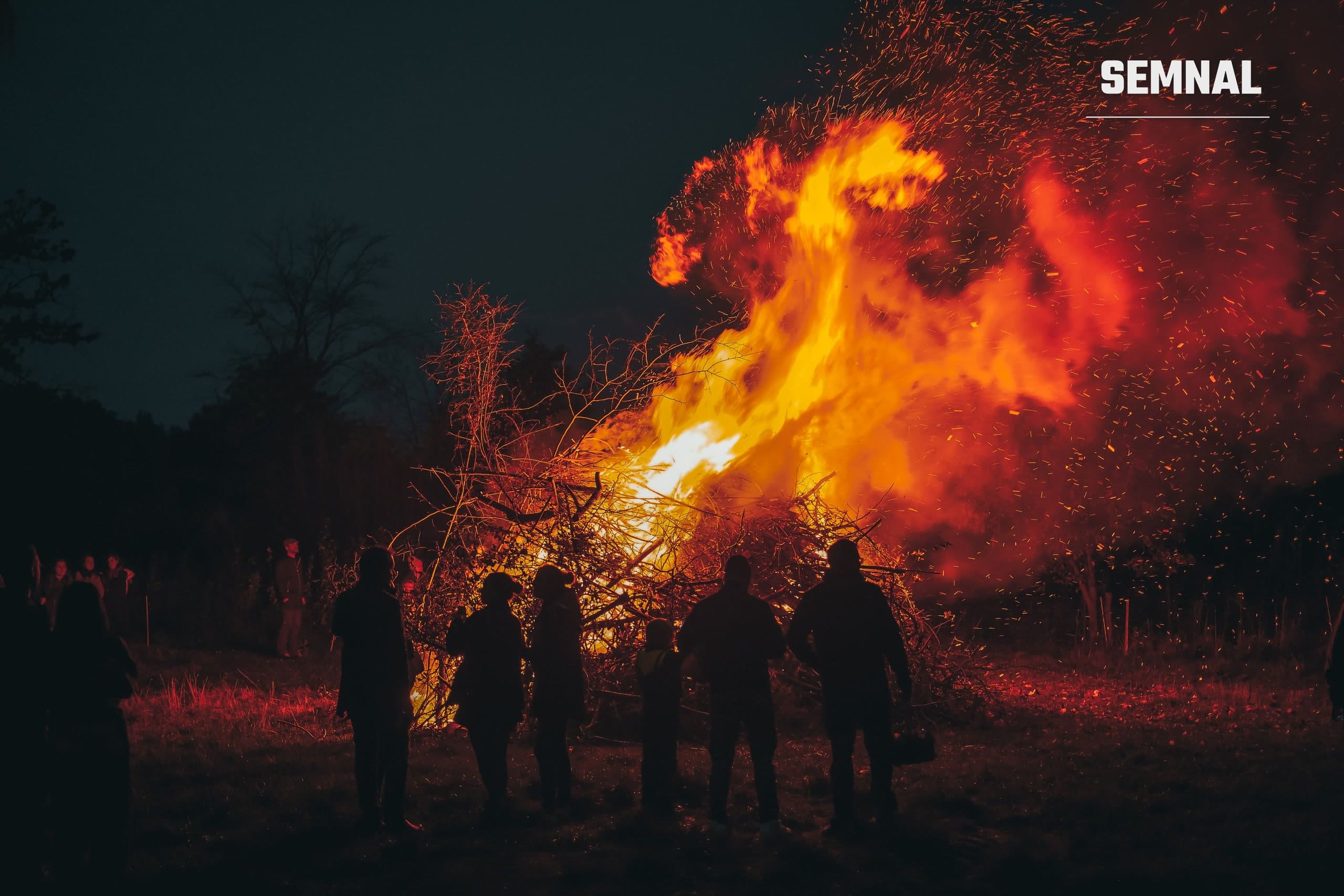 Siluete ale unor oameni în fața unui rug mare aprins noaptea, cu flăcări și scântei, fotografie ilustrativă
