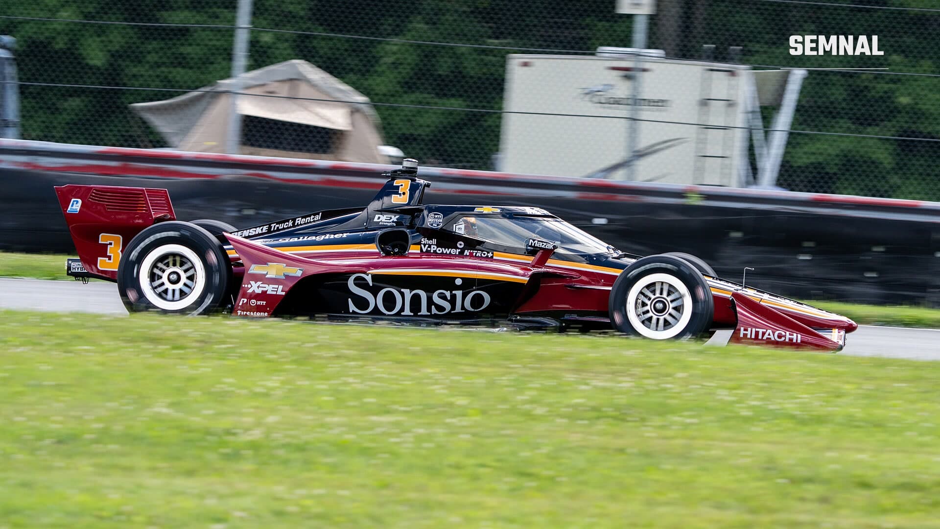 Monopost IndyCar roșu-vișiniu cu negru, cu numărul 3, în viteză pe un circuit rutier, cu iarbă în prim-plan și bariera de protecție în fundal
