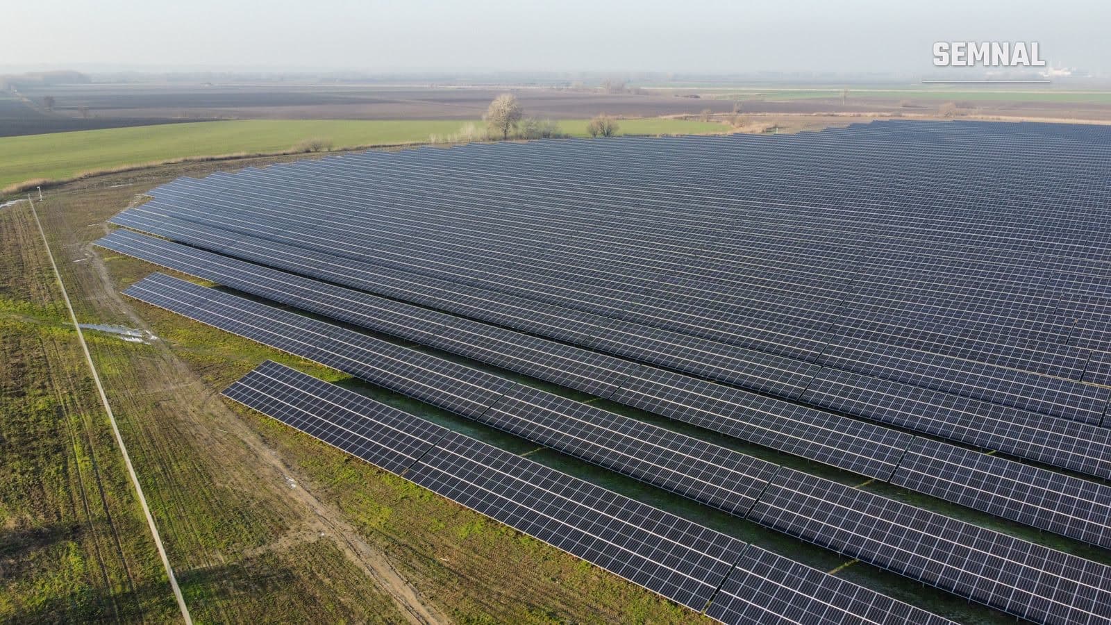 Vedere aeriană a unui parc fotovoltaic întins, cu rânduri lungi de panouri solare pe un teren agricol plat, într-o zi cu ușoară ceață