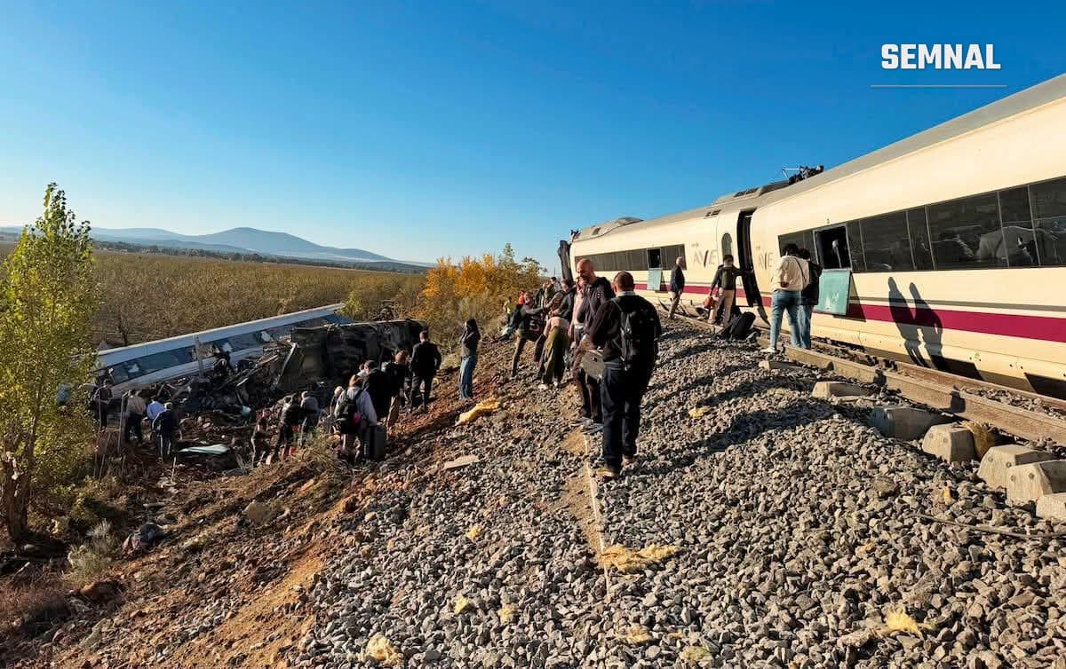 Pasageri merg pe lângă un tren AVE oprit pe calea ferată, iar în stânga se văd resturile unui vagon căzut pe taluz