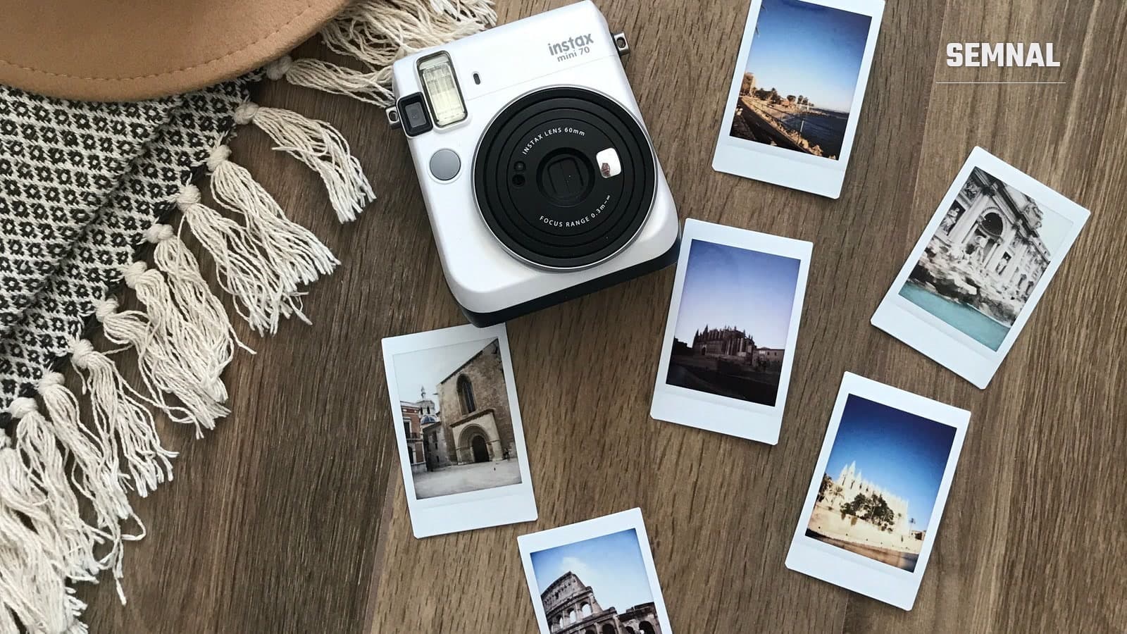 Aparat foto instant alb și șase fotografii tipărite cu clădiri istorice, așezate pe un parchet de lemn, lângă o pălărie și un covor cu franjuri
