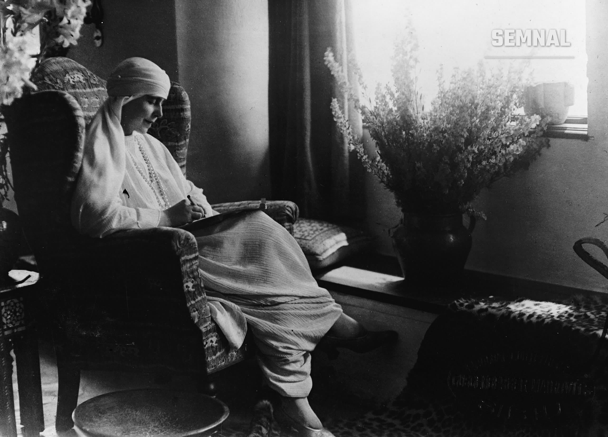 Regina Maria a României, într-o fotografie alb-negru de epocă, scrie într-un fotoliu, lângă o vază mare cu flori, la o fereastră.