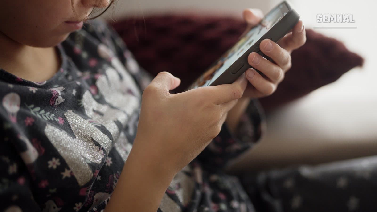 Copil care folosește un telefon mobil, așezat pe o canapea, cu fața parțial ascunsă de cadru