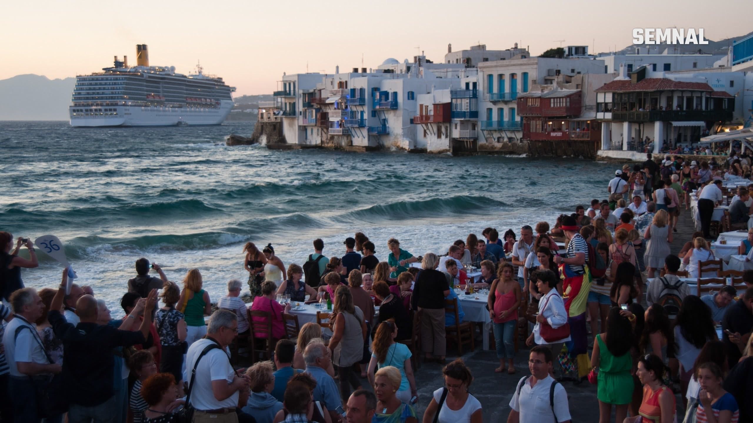 Turiști la mese și pe chei, la apus, în cartierul Little Venice din Mykonos, cu un vas de croazieră pe mare în fundal