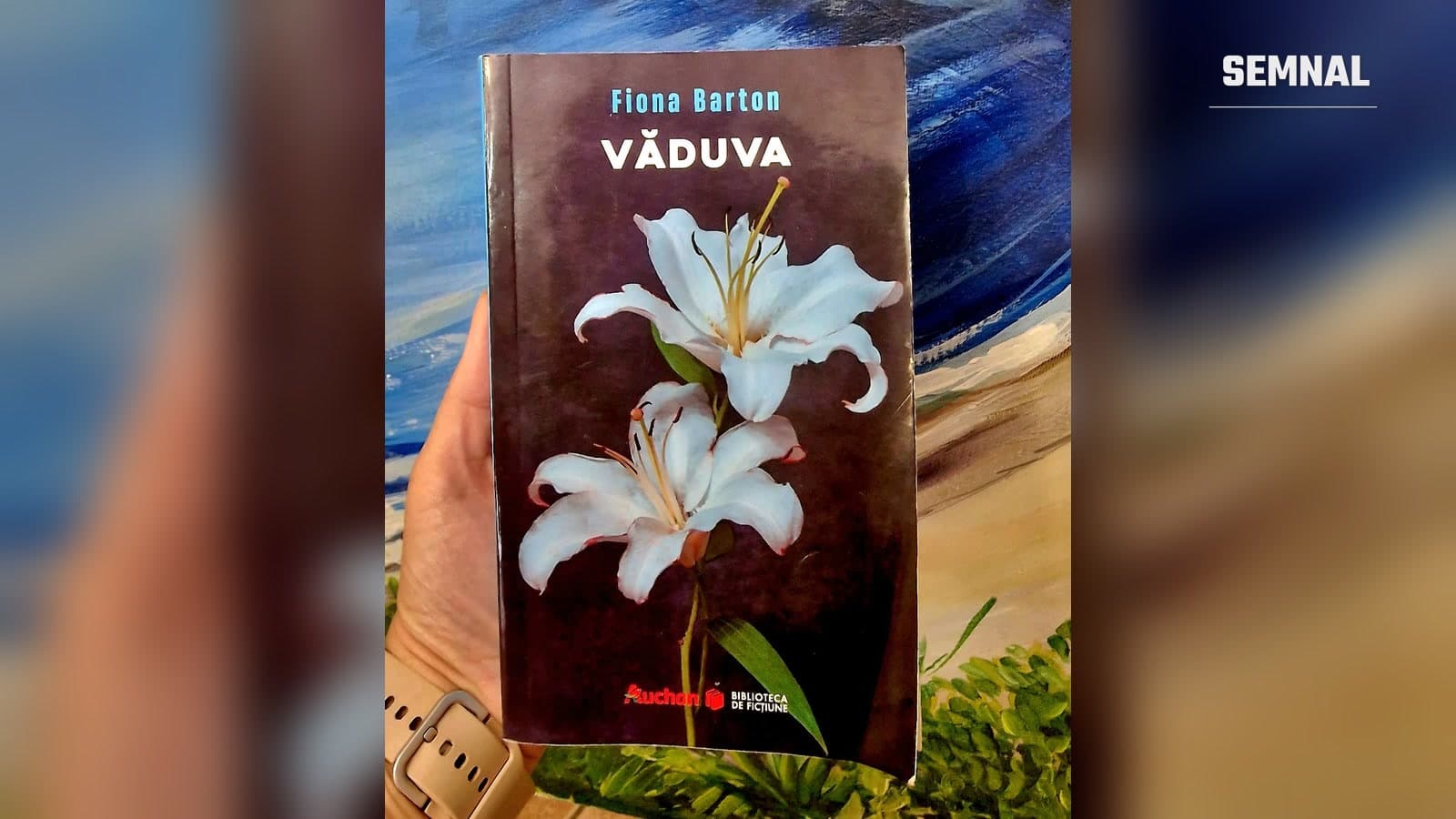 Romanul Văduva de Fiona Barton, ediția Biblioteca de ficțiune Auchan, ținut în mână în fața unui tablou cu o mare albastră