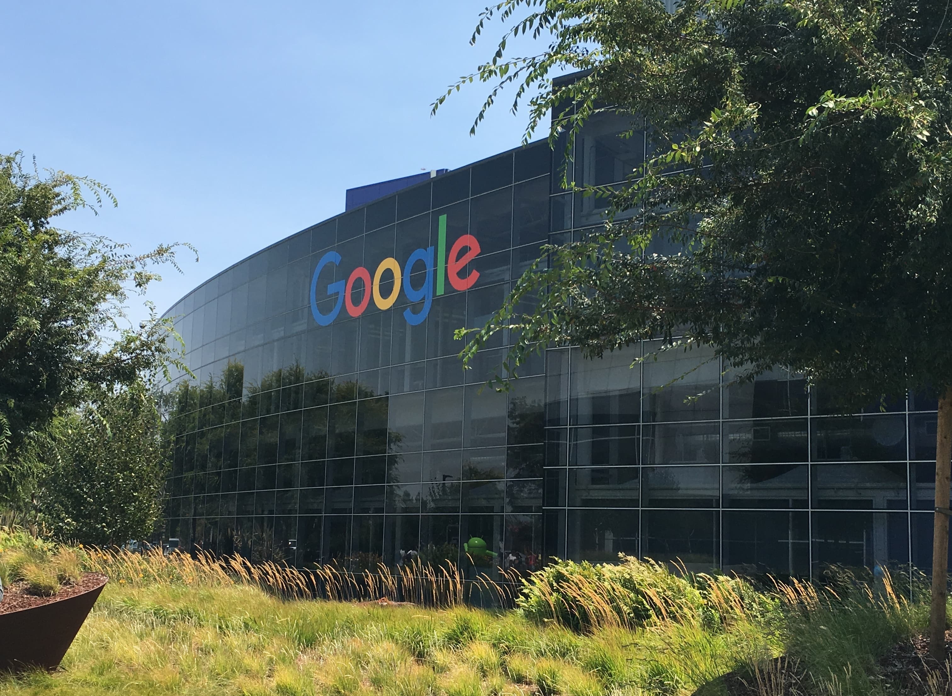 Fațada modernă a clădirii Google cu logo-ul multicolor pe o clădire cu pereți de sticlă neagră, înconjurată de copaci și vegetație verde pe o zi însorită.