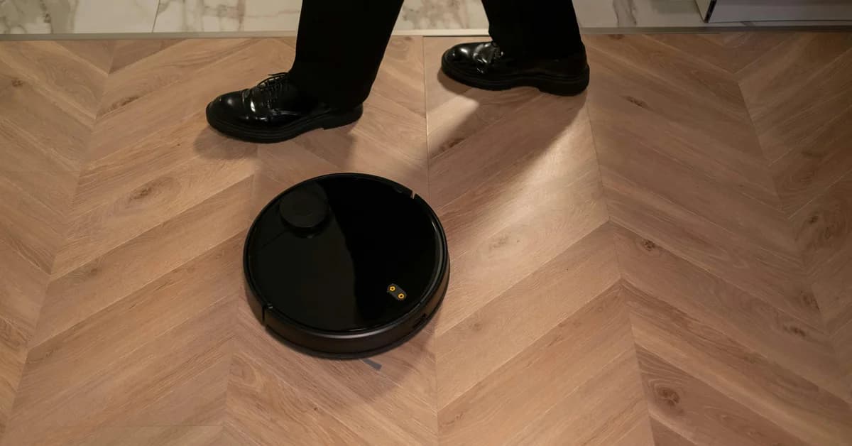 Un robot de curățenie Deebot X12 se deplasează pe un parchet în model chevron, lângă picioarele unei persoane.