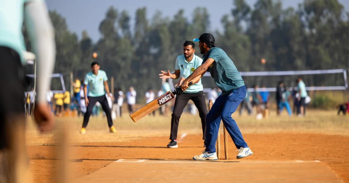 Jucători de cricket în echipament albastru, în timpul unui meci desfășurat pe un teren cu iarbă galbenă, pe fundal fiind alți jucători și spectatori.