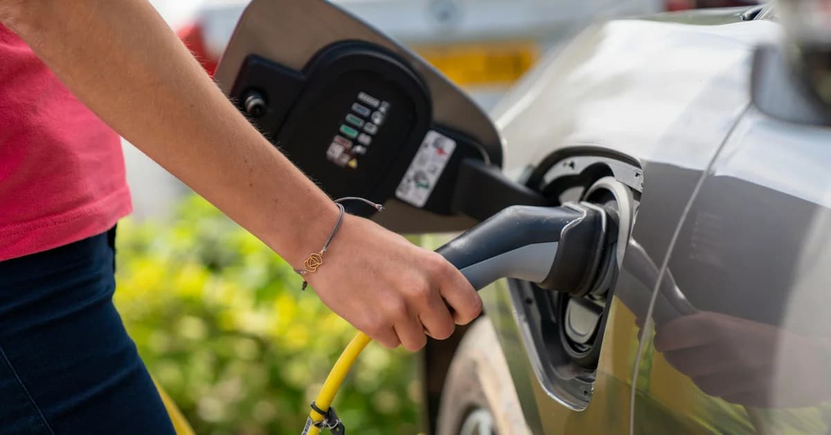 O persoană conectează un cablu la portul de încărcare al unei mașini electrice, în fundal alte mașini parcate.