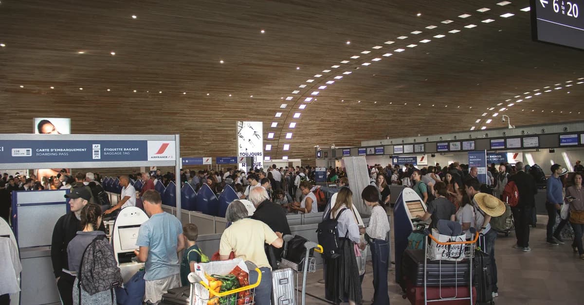 Pasageri așteaptă la controlul de securitate într-un aeroport aglomerat cu tavan curbat și panouri de afișaj electronic.