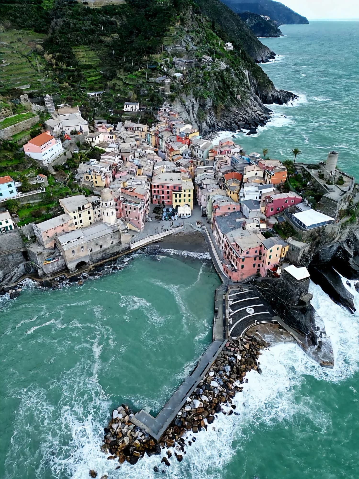Vedere aeriană a unui orășel pescăresc italian cu case colorate pe stâncă, port mic și mare turcoaz, peisaj montan verde în fundal.
