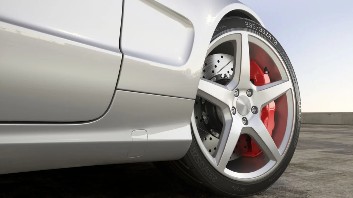 Detaliu al roții unei mașini cu jantă sport alb-argintie, discuri de frână gri și piston de frână roșu, ilustrând sistemul de frânare al unui automobil.