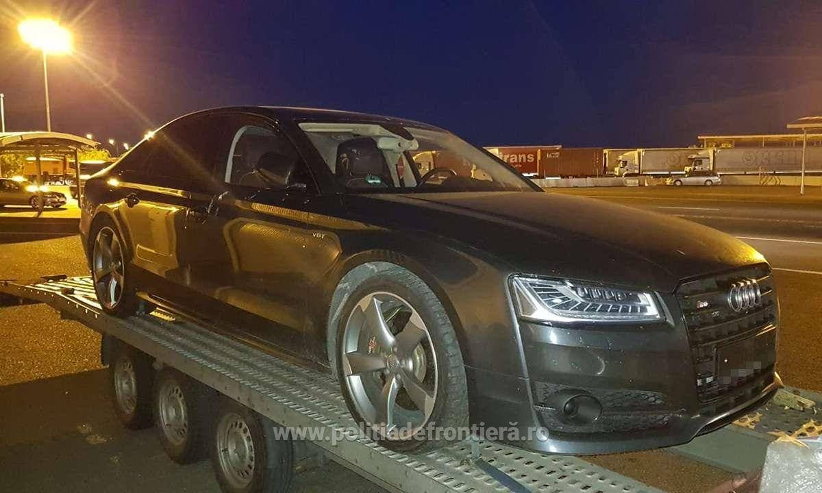 Autoturism negru de lux Audi transportat pe remorcă de transport, noaptea, în zona de terminal cu containere, ilustrând logistica aducerii unui automobil din Uniunea Europeană.